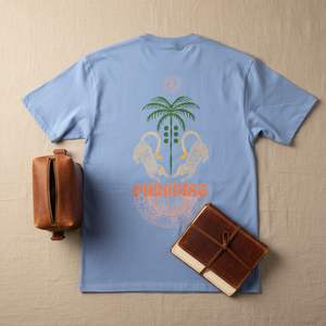 Blue t-shirt with a palm tree and flamingo design on a beige surface with a leather bag and book.